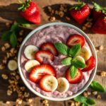 Creamy smoothie topped with strawberries, bananas, mint leaves, and granola on a wooden table